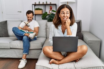 Poster - Hispanic middle age couple at home, woman using laptop looking stressed and nervous with hands on mouth biting nails. anxiety problem.