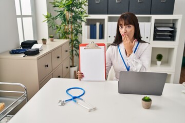 Poster - Young brunette doctor woman holding clipboard covering mouth with hand, shocked and afraid for mistake. surprised expression