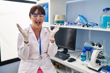 Canvas Print - Young brunette woman working at scientist laboratory celebrating mad and crazy for success with arms raised and closed eyes screaming excited. winner concept