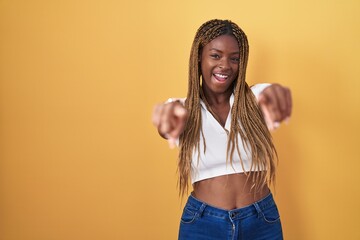 Wall Mural - African american woman with braided hair standing over yellow background pointing to you and the camera with fingers, smiling positive and cheerful