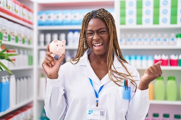 Canvas Print - African american woman with braided hair working at pharmacy drugstore holding piggy bank screaming proud, celebrating victory and success very excited with raised arm