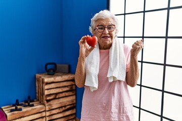 Wall Mural - Senior woman with grey hair wearing sportswear holding red heart screaming proud, celebrating victory and success very excited with raised arm