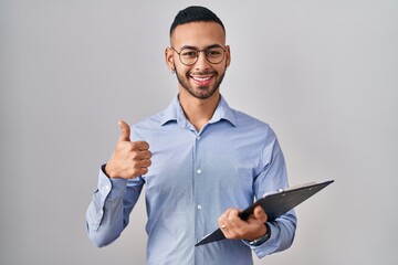 Wall Mural - Young hispanic man wearing business clothes holding clipboard smiling happy and positive, thumb up doing excellent and approval sign