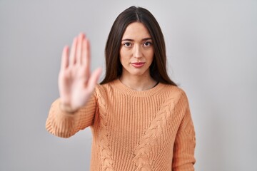 Sticker - Young brunette woman standing over white background doing stop sing with palm of the hand. warning expression with negative and serious gesture on the face.