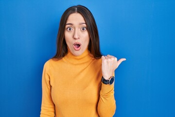 Sticker - Young brunette woman standing over blue background surprised pointing with hand finger to the side, open mouth amazed expression.