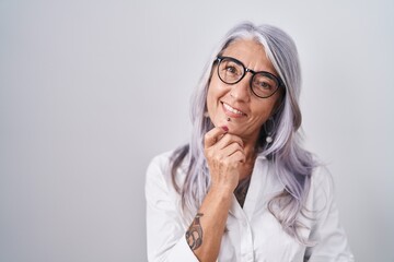 Poster - Middle age woman with tattoos wearing glasses standing over white background looking confident at the camera smiling with crossed arms and hand raised on chin. thinking positive.