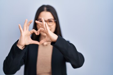 Poster - Young brunette woman standing over blue background smiling in love doing heart symbol shape with hands. romantic concept.