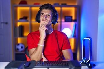 Wall Mural - Young hispanic man playing video games with hand on chin thinking about question, pensive expression. smiling with thoughtful face. doubt concept.