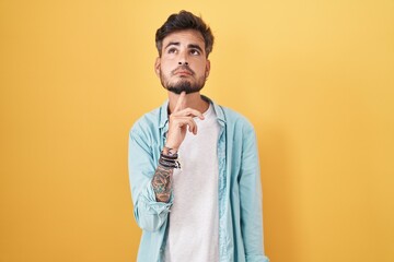 Wall Mural - Young hispanic man with tattoos standing over yellow background thinking concentrated about doubt with finger on chin and looking up wondering