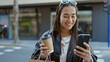 © Krakenimages.com - Young beautiful hispanic woman using smartphone holding shopping bags and coffee at street
