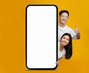 Wall Mural - Online Offer. Smiling Asian Couple Peeking Out Behind Big Blank Smartphone