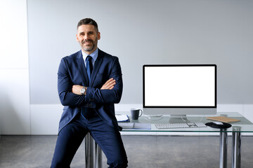 Poster - Confident middle aged businessman leaning on table, posing with folded arms near computer with blank screen, mockup