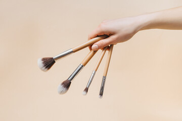 Female fragile hand holding makeup brushes with wooden base on beige isolated background. Concept of natural cosmetic products for skin care, beauty and fashion