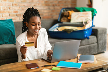 happy black woman using laptop and booking tour online with credit card, sitting with unpacked suitc
