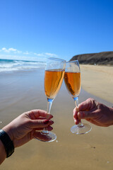 Wall Mural - Beach party, hands with glass of rose champagne or cava sparkling wine, view on white sandy tropical beach and blue ocean water, romantic vacation, winter sun on Fuerteventura, Canary, Spain