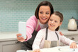 © Prostock-studio - Smiling caucasian young mother and little daughter in aprons take selfie on phone, make cookies in kitchen
