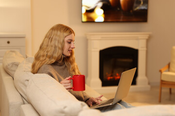 Poster - Young woman with cup of hot drink working on laptop near fireplace at home