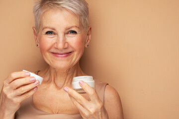 Smiling 60s middle aged mature woman putting tightening facial cream on face looking at camera.