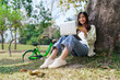 © snowing12 - Women sitting under tree and holding credit card to shopping online on laptop after cycling in park