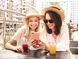 © halayalex - Two young beautiful smiling hipster female in trendy summer white dress and hats. Sexy carefree women drinking fresh vegetable cocktail smoothie drink in plastic cup with straw. Using phone apps