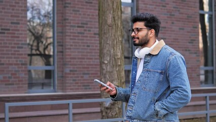 A good looking Latino man with headphones walks down the street, chatting on his mobile phone, typing a message while scrolling. Surfing social media, enjoying his free time and the holiday concept.