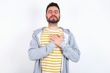 Wall Mural - Young caucasian mán wearing trendy clothes over white background smiling with hands on chest with closed eyes and grateful gesture on face. Health concept.