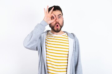 Wall Mural - Young caucasian mán wearing trendy clothes over white background doing ok gesture shocked with surprised face, eye looking through fingers. Unbelieving expression.