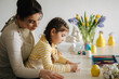 © Aleksandr - Side view of mom helping her daughter paint an Easter bunny mask. Little girl sitting at the table in the kitchen