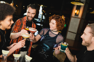 Wall Mural - overjoyed multiethnic friends holding glasses with cocktails near blurred tequila shots in bar.