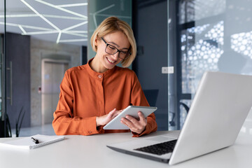 Young successful businesswoman at workplace is satisfied with results of achievement at work smiling and using tablet computer, blonde programmer is testing new software in application.