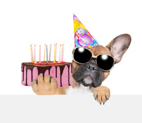 Poster - French bulldog puppy wearing sunglasses and party cap holding birthday cake with many candles looking above empty banner. isolated on white background