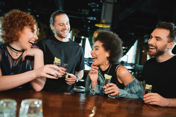 Wall Mural - Carefree interracial people holding tequila shots near stand in bar.