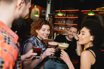 Wall Mural - Smiling multiethnic women clinking cocktails near blurred friends in bar.