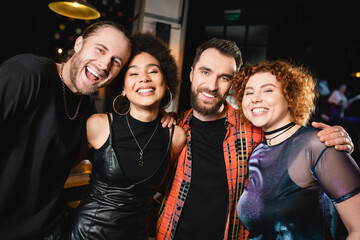 Wall Mural - Smiling multiethnic friends hugging and looking at camera in bar in evening.