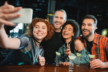 Wall Mural - Smiling multiethnic friends taking selfie on smartphone near tequila in bar.