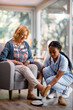 Young black nurse helps senior woman to take off her shoes in nursing home.