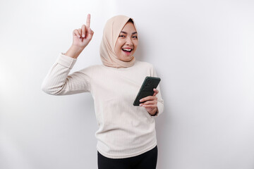 Excited Asian woman wearing hijab pointing at the copy space on top of her while holding her phone, isolated by white background