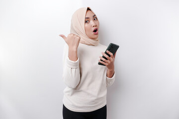 Shocked Asian Muslim woman wearing hijab pointing at the copy space beside her while holding her phone, isolated by white background