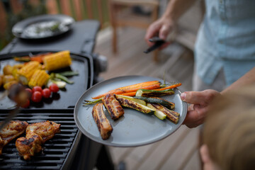 Wall Mural - Father putting grilled meat and vegetable on plate to his son during family summer garden party.