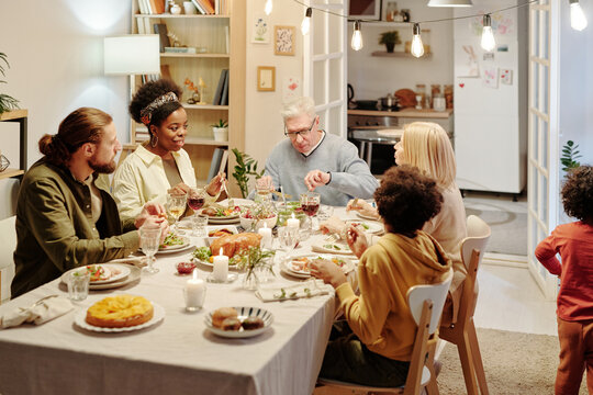 Members of large intercultural family sitting and communicating by served table while eating homemade food at dinner in living room