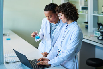 Poster - Two intercultural female virologists studying liquid sample in test tube and searching online information describing its characteristics