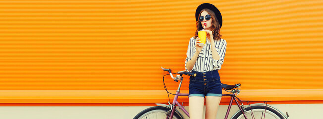 Summer colorful image of happy young woman drinking fresh juice with bicycle looking away on orange background