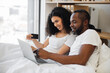 © sofiko14 - Loving multiethnic family spending morning in bed with laptop while using credit card for internet banking at home. Romantic partners making online payment operations in available application.