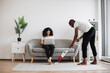 © sofiko14 - Side view of african man cleaning living room with cordless vacuum while focused woman working on laptop on couch. Caring husband doing households while wife freelancing at home.