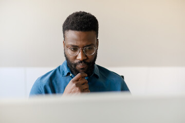 Focused african american male ceo touching chin and looking at computer screen, working at desk in office, copy space