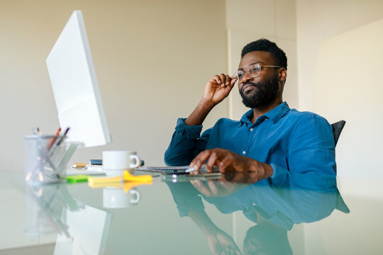 Wall Mural - Black businessman sitting at desk in office and working on computer, handsome male entrepreneur reading corporate emails