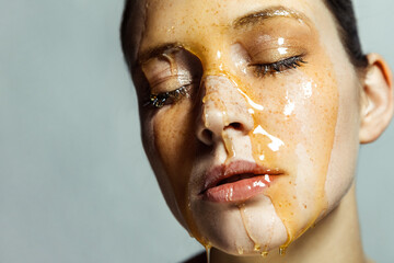 Attractive calm woman with freckles and honey on face, keeps eyes closed, having serious expression.