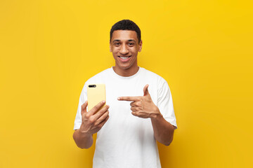 Wall Mural - young african american guy in white t-shirt points finger at smartphone on yellow isolated background