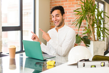 Wall Mural - young hispanic man smiling cheerfully and pointing to copy space on palm on the side, showing or advertising an object. architect concept