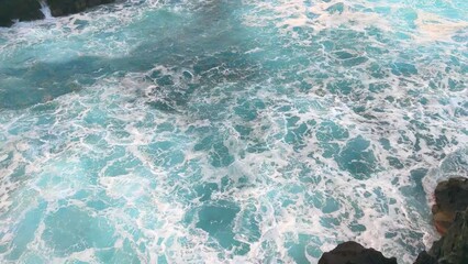 Wall Mural - Turquoise waves and foam crash against the rocks. View from a height